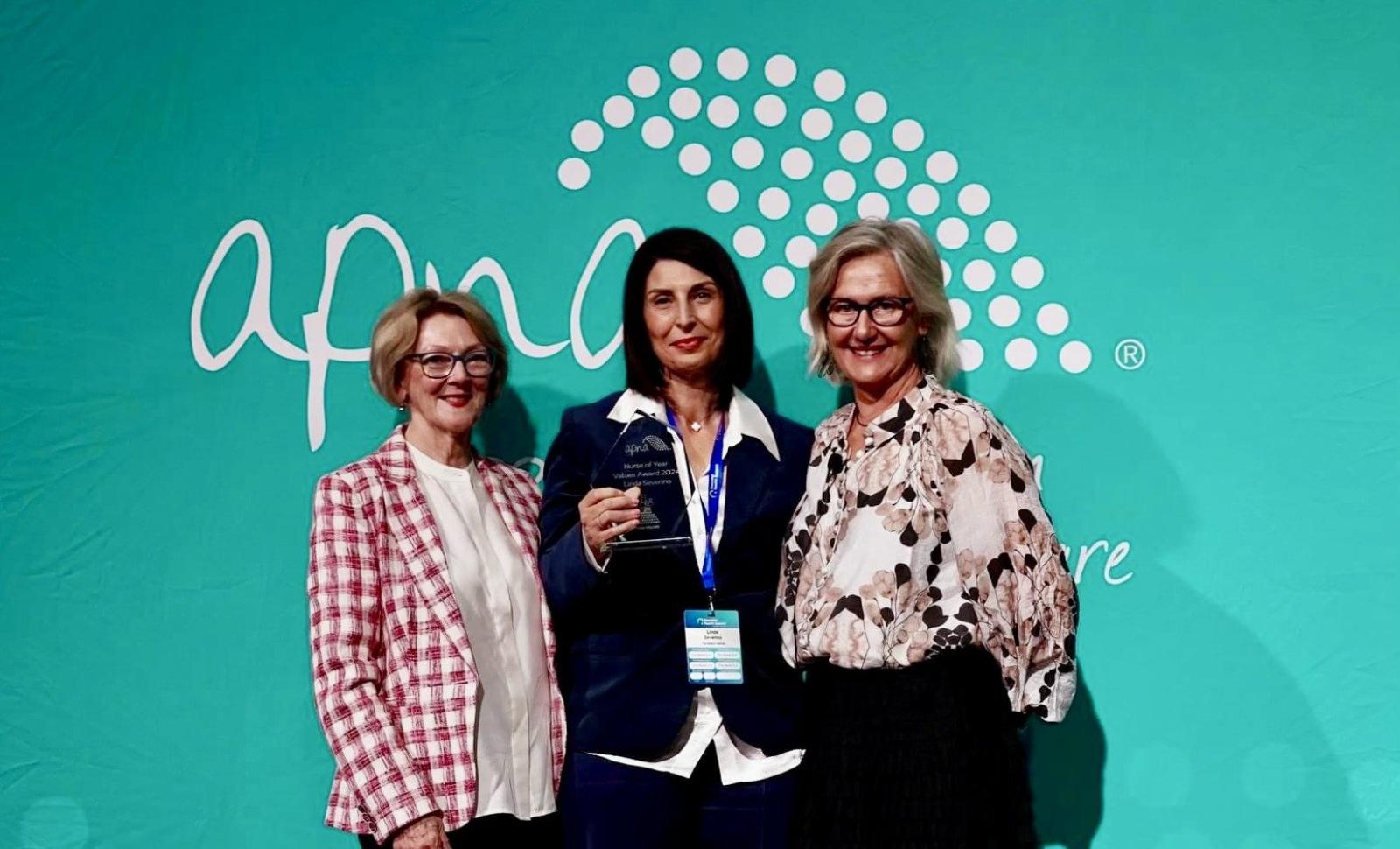Three women pose at a healthcare awards event in front of a teal Apicare backdrop